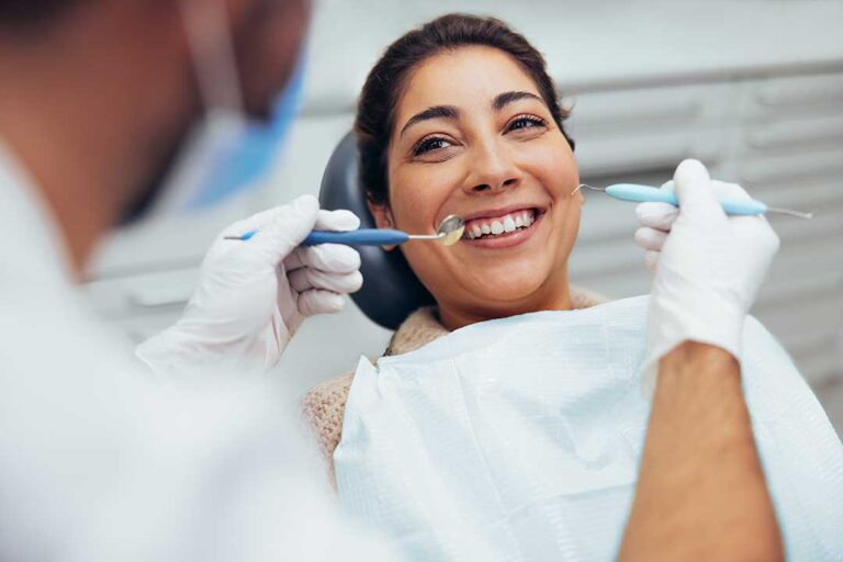 Person gently brushing near the gumline beside a glass of water and a tube of sensitive-teeth toothpaste.